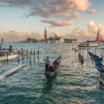 Iconic view of San Giorgio Maggiore church and traditional Venetian gondolas with tourists during sunset in Venice, Italy.
