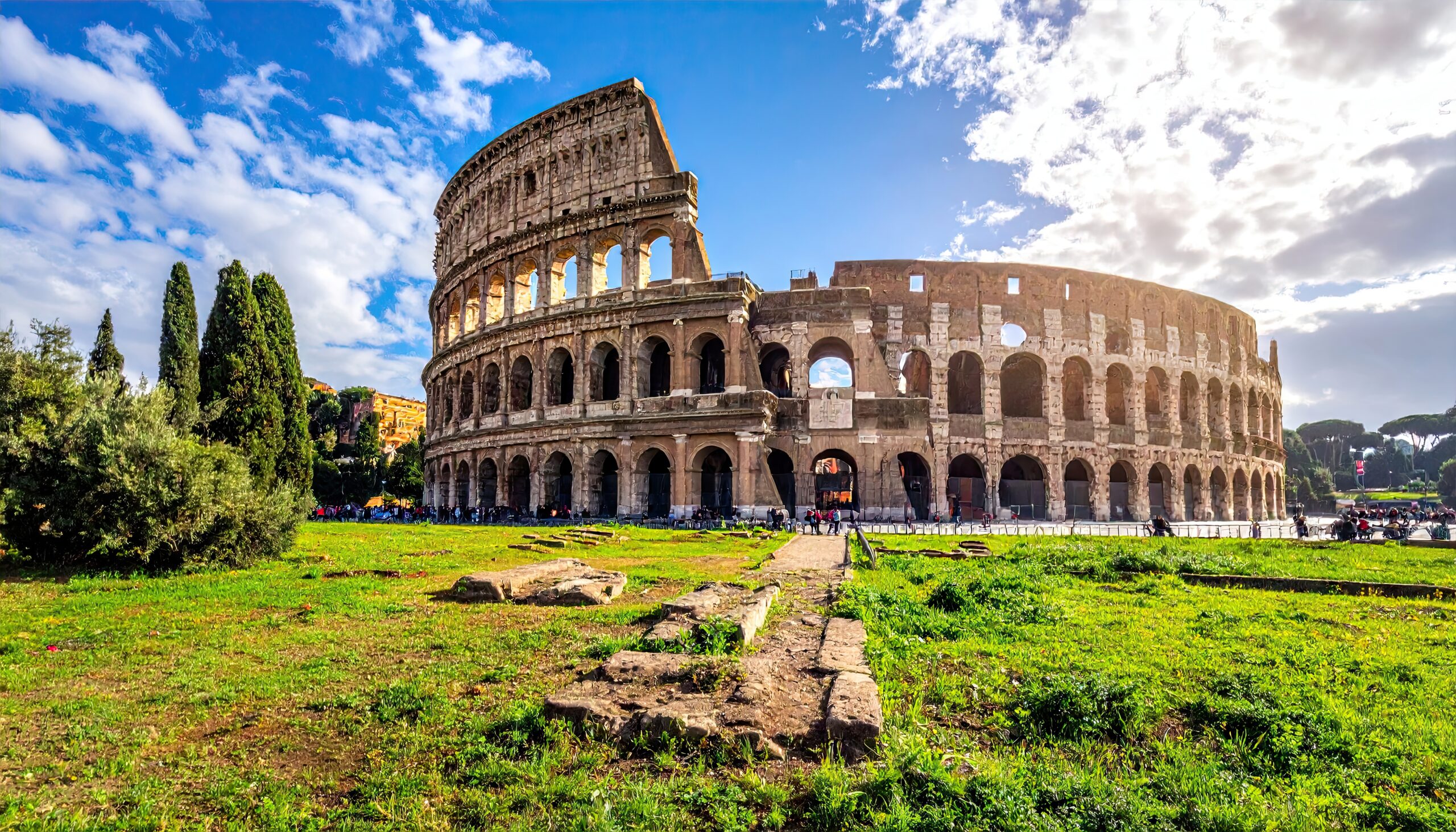 Ancient Roman Colosseum Amphitheater Iconic Landmark Under Sunny Blue Sky With Green Grass And Trees In Rome Italy